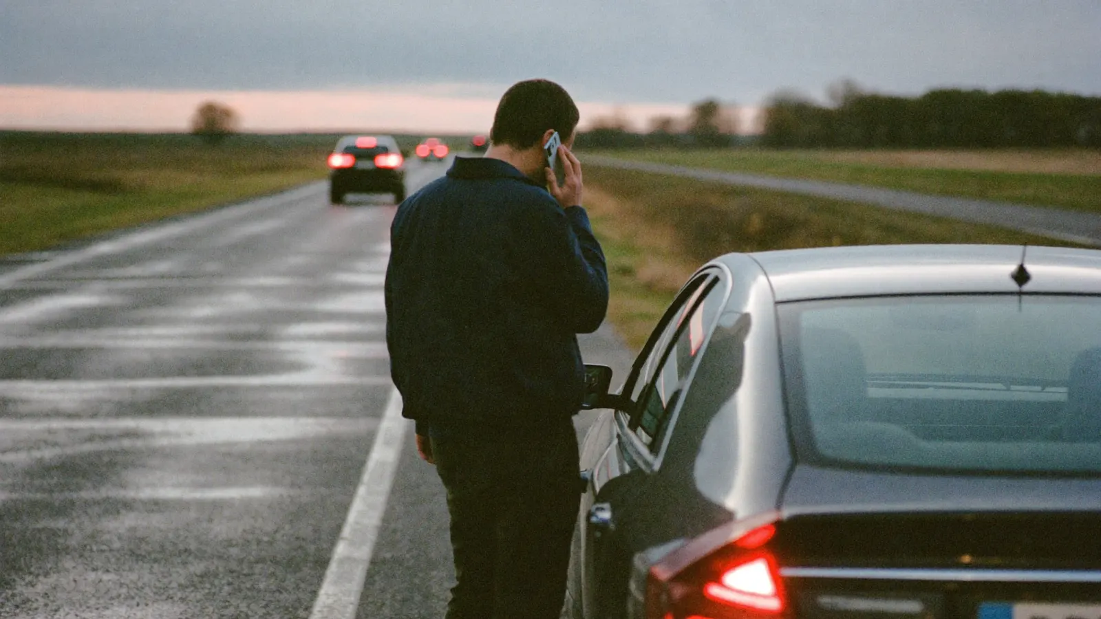 Een man staat in de schemer langs een natte provinciale weg naast zijn auto, met een telefoon aan zijn oor.